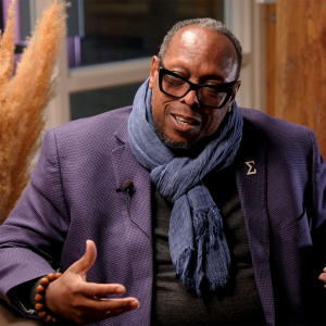 Series host Mark Anthony Neal, an African American man in his fifties, dressed in a blue suit jacket with a blue scarf and a black shirt, wearing glasses