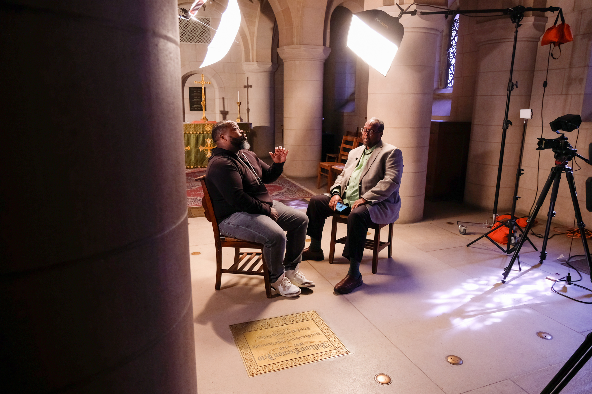 two men sit in a make-shift studio speaking to each other